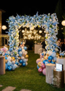 An 8ft x 8ft Yellow White Rose Roll Up Flower Backdrop with fabric backing creates a radiant floral arch cover, forming an elegant setting for weddings, parties, or ceremonies outdoors at night.