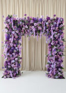 The Burgundy Red Rose Flower Arch Backdrop (8ft x 8ft) features lush purple and lavender flowers before cream drapes and a burgundy background on a white floor, creating an elegant, romantic wedding display.