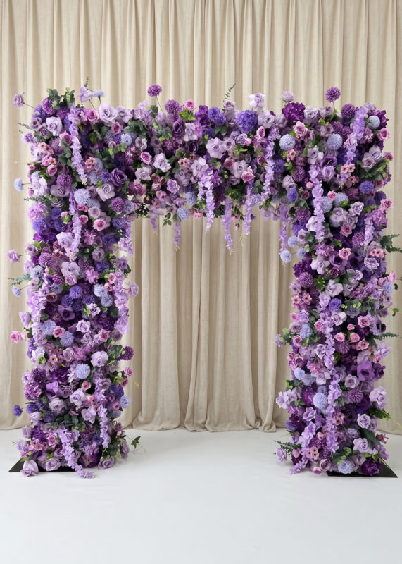 The Burgundy Red Rose Flower Arch Backdrop (8ft x 8ft) features lush purple and lavender flowers before cream drapes and a burgundy background on a white floor, creating an elegant, romantic wedding display.