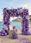 The Burgundy Red Rose Flower Arch Backdrop (8ft x 8ft) creates a stunning romantic setting by the sea, with boats and a coastal town behind. Two pedestals display a sign and bouquet, while petals are scattered on the stone ground.