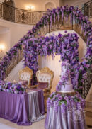 Elegant wedding setup with gold-trimmed chairs, Burgundy Red Rose Flower Arch Backdrop (8ft x 8ft), draped staircase, satin tablecloth, and a tiered purple cake with cascading flowers. Luxurious, romantic indoor venue.