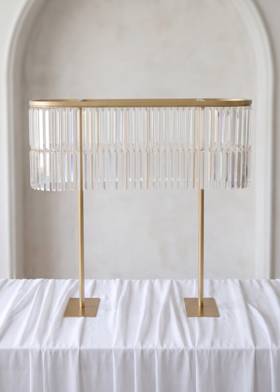 A rectangular gold metal centerpiece stand draped with hanging crystal prisms, standing on a white tablecloth.