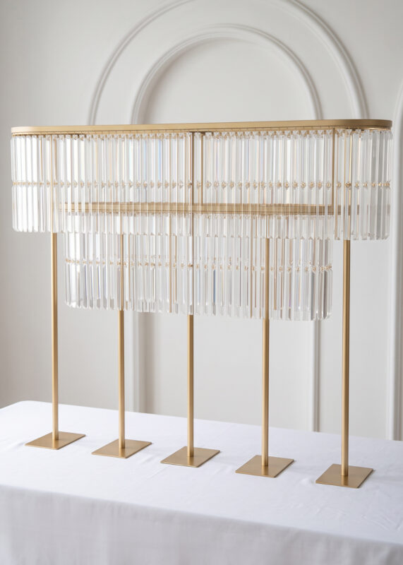 A 51.2in Gold Double-Layer Crystal Fringe Wedding Centerpiece Stand with an oval design, featuring hanging prisms and five vertical support posts on a white tablecloth.