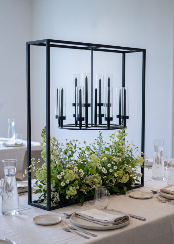 A 39.4in Black Square Frame Hanging Candelabra Centerpiece featuring a 12-arm candle holder above a floral arrangement of white daisies and green foliage on a set dining table.