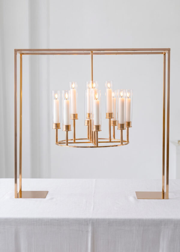 A 39.4in Black Square Frame Hanging Candelabra Centerpiece in metallic gold-tone with 12 lit candles in glass sleeves, centered on a white tablecloth.