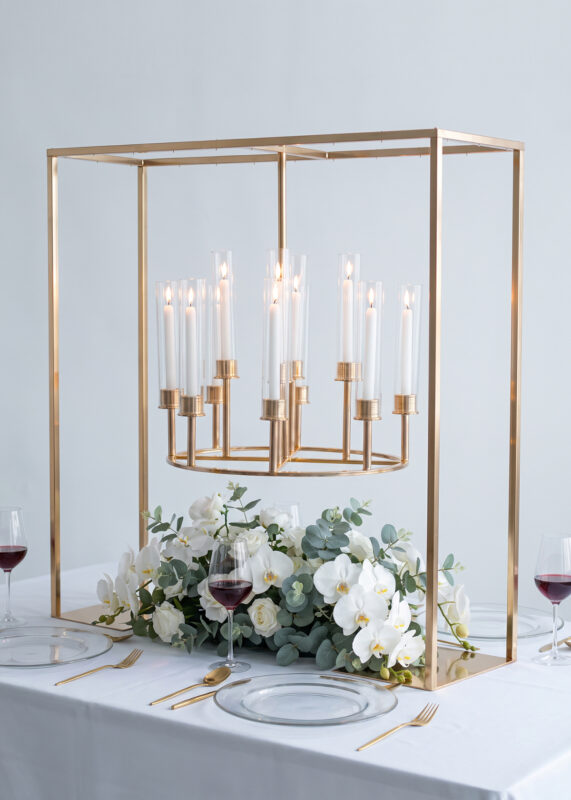 A 12-Arm Gold Hanging Candelabra Centerpiece, Luxurious & Romantic 39in Square Wedding Candle Stand holds a circular, twelve-arm frame with lit taper candles, positioned over white orchids and greenery on a set dining table.