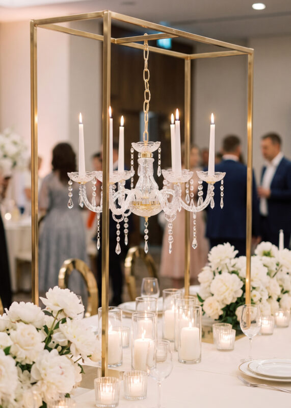 A gold Wedding Table Hanging Acrylic Chandelier Stand with a square frame and six candle bowls sits on a dining table, holding a crystal fixture with lit taper candles surrounded by white flower arrangements and glass candle holders.