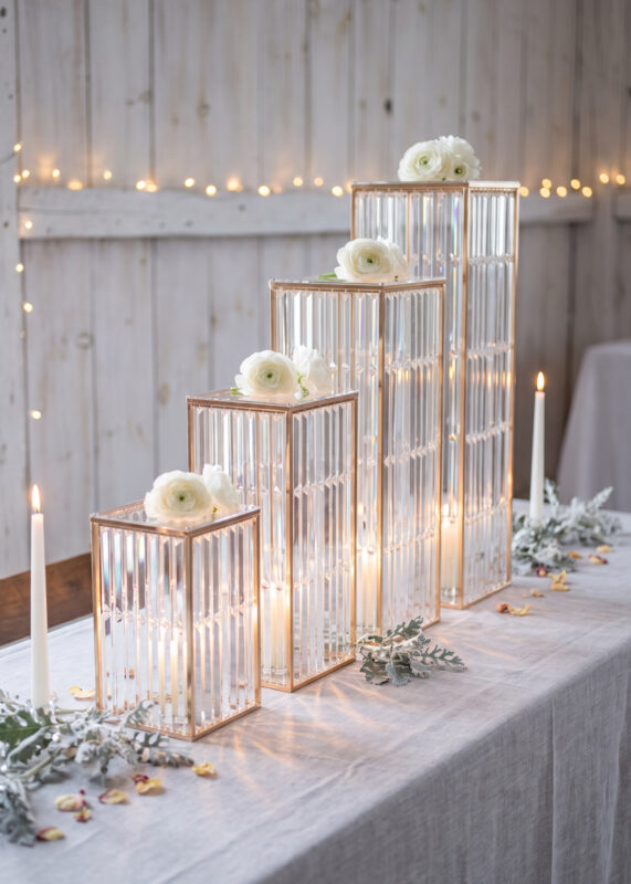 A set of 4 Gold Crystal Stripe Pedestal Stands of varying heights arranged in a row on a table, each topped with a white flower and containing a lit candle.